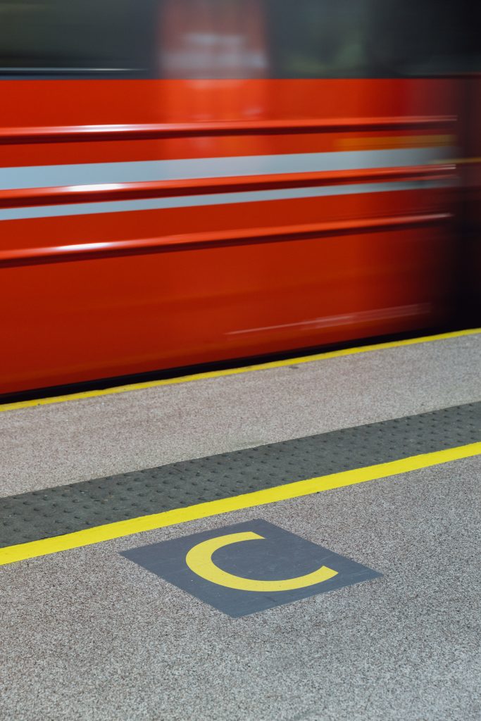 Grey anti-slip floor coating with yellow marking lines on a railway platform, a red train speeding past behind