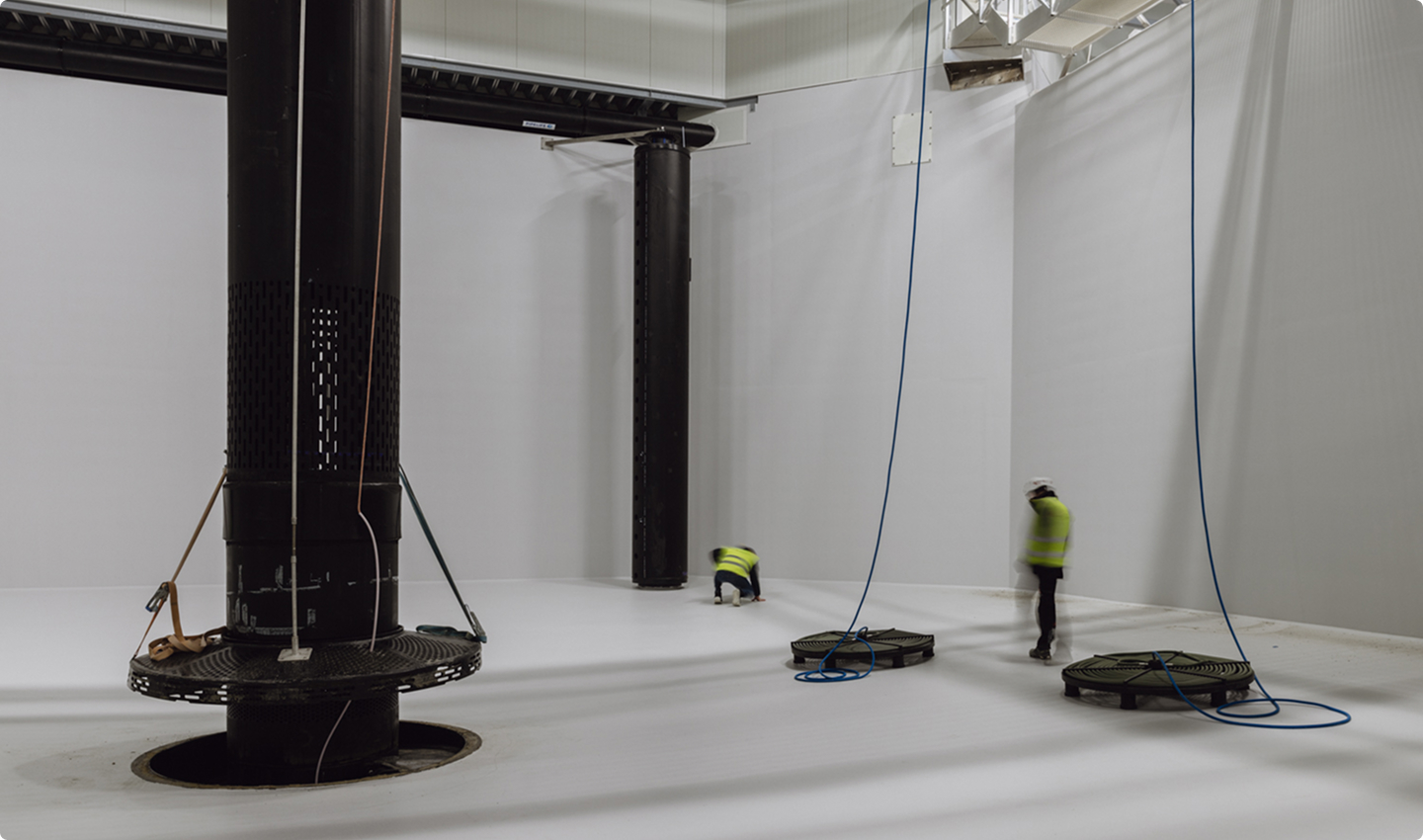 Two workers in hi-vis vests on light industrial flooring in a large process hall with black pipe columns